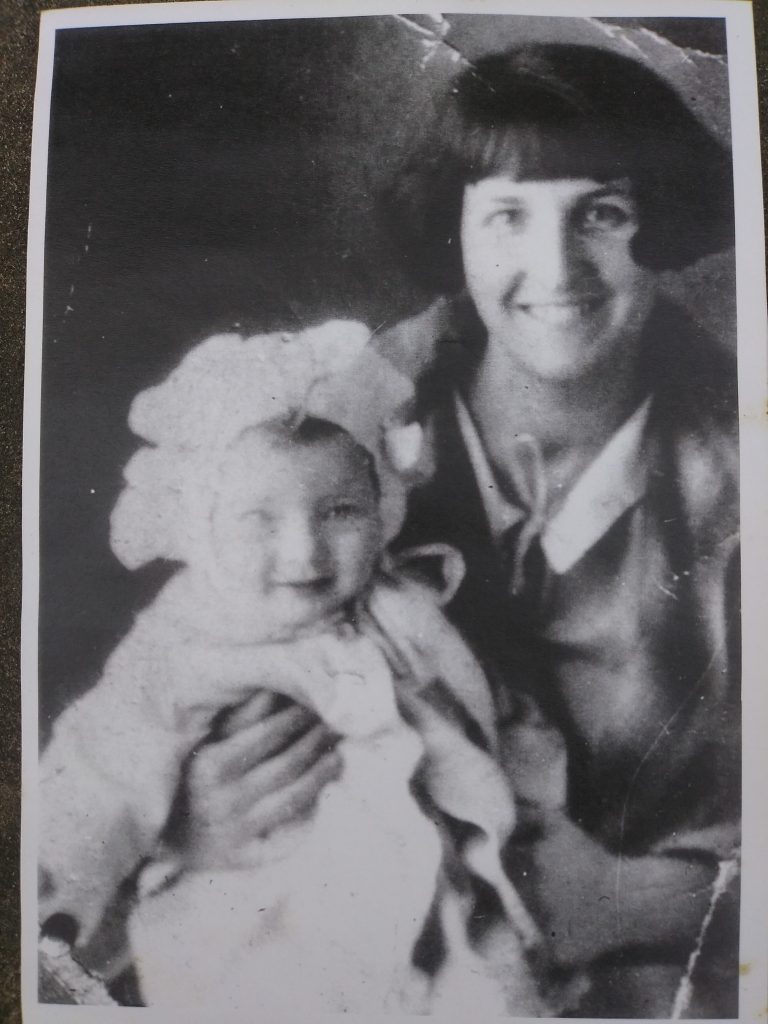 Black and White photo of a baby wearing a bonnet sitting on the lap of a woman with dark hair cut in a short bob. Both are smiling. 